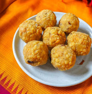 Six golden-brown fried sweet balls, likely laddoos or similar Indian sweets, arranged on a small white plate over a bright orange patterned fabric background.