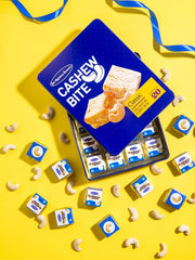 A tin of Classic Cashew Bites on a bright yellow background, surrounded by loose cashews and smaller wrapped pieces. The tin is dark blue with white and yellow text and images of cashew pieces.