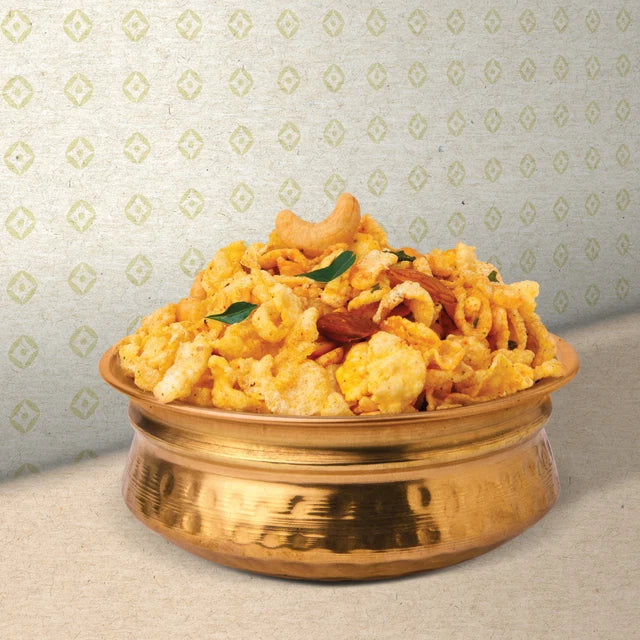 A bowl of golden fried snack pieces, likely Indian chivda or sambar, garnished with cashews and green leaves, served in a metallic brass bowl against a patterned light gray background.