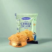 A bag of Sri Krishna Swastey Green Chilli Blood Thhattai snack stands behind two copper bowls filled with the golden-brown fried snacks, set against a solid sage green background.