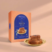 A box of St. Kitchen Brothers Royal Maha Mittai next to a small gold plate holding two pieces of the brown, dense sweet snack, set against a warm beige background with soft shadows.