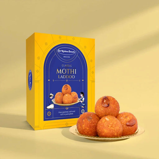 A yellow and blue box of St. Kitchen Brothers Classic Mothi Laddoo stands next to a small plate of golden-brown laddoo pieces on a warm beige background.