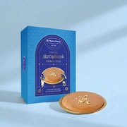 A blue boxed dessert product labeled 'Classic Therattipaal' stands next to a round golden-brown baked good on a small gold plate, photographed on a clean light blue studio background.